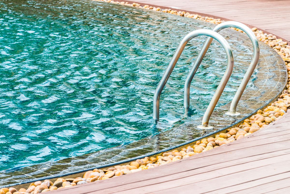 Dettaglio di piscina con scala in acciaio inox, bordo curvo con sassi decorativi e pavimentazione in legno, elementi e accessori della piscina